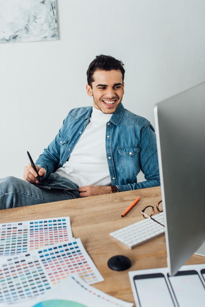 Glimlachende ux ontwerper met behulp van grafische tablet en computer in de buurt van kleurenpaletten op tafel  - Foto, afbeelding