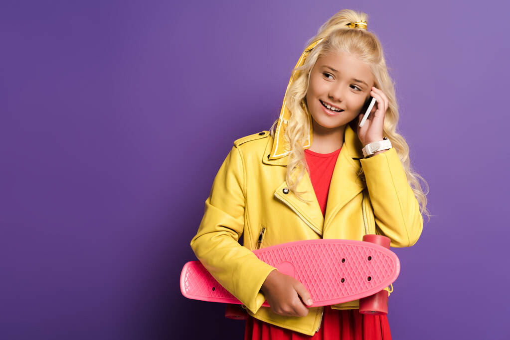 glimlachend kind houden penny board en praten op smartphone op paarse achtergrond  - Foto, afbeelding