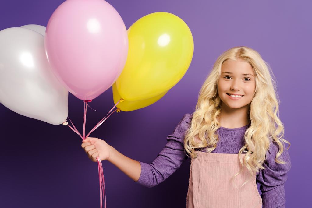 niño sonriente sosteniendo globos y mirando a la cámara sobre fondo púrpura
  - Foto, imagen