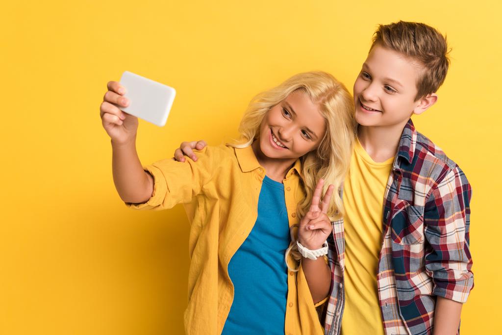 Lächelndes Kind zeigt Friedensgeste und macht Selfie mit Freund auf gelbem Hintergrund  - Foto, Bild