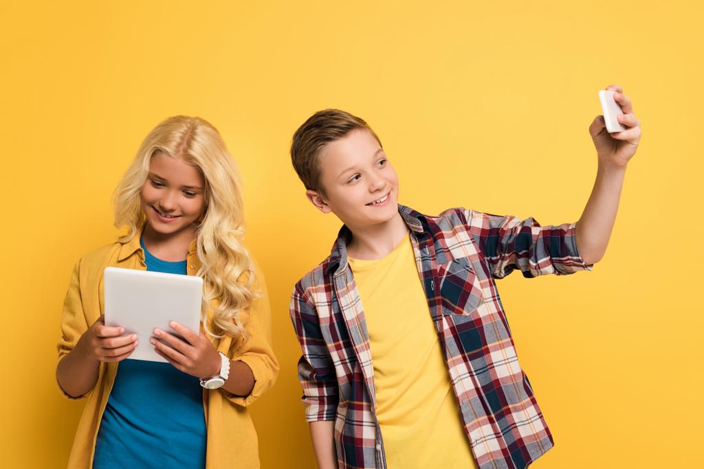 lachend kind het nemen van selfie en zijn vriend met behulp van digitale tablet op gele achtergrond  - Foto, afbeelding