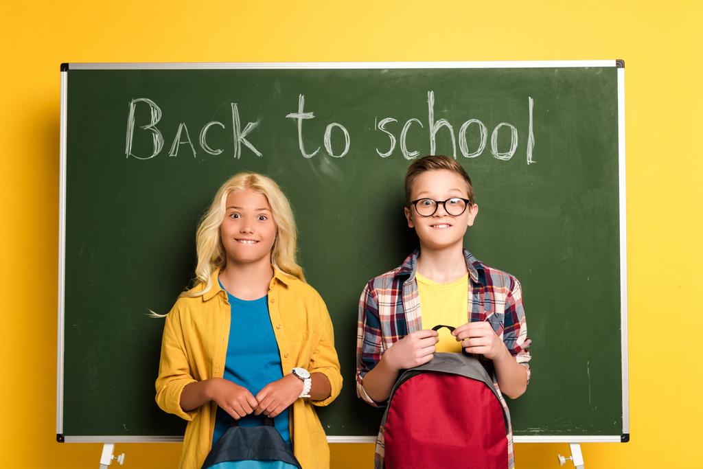 crianças em idade escolar sorridentes com mochilas em pé perto de quadro-negro com letras de volta à escola
  - Foto, Imagem