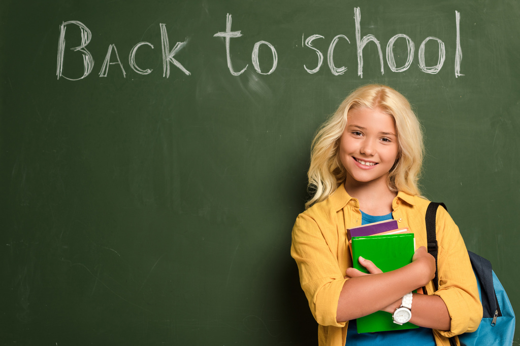 χαμογελαστή μαθήτρια με βιβλία που στέκονται κοντά στο chalkboard με γράμματα πίσω στο σχολείο - Φωτογραφία, εικόνα