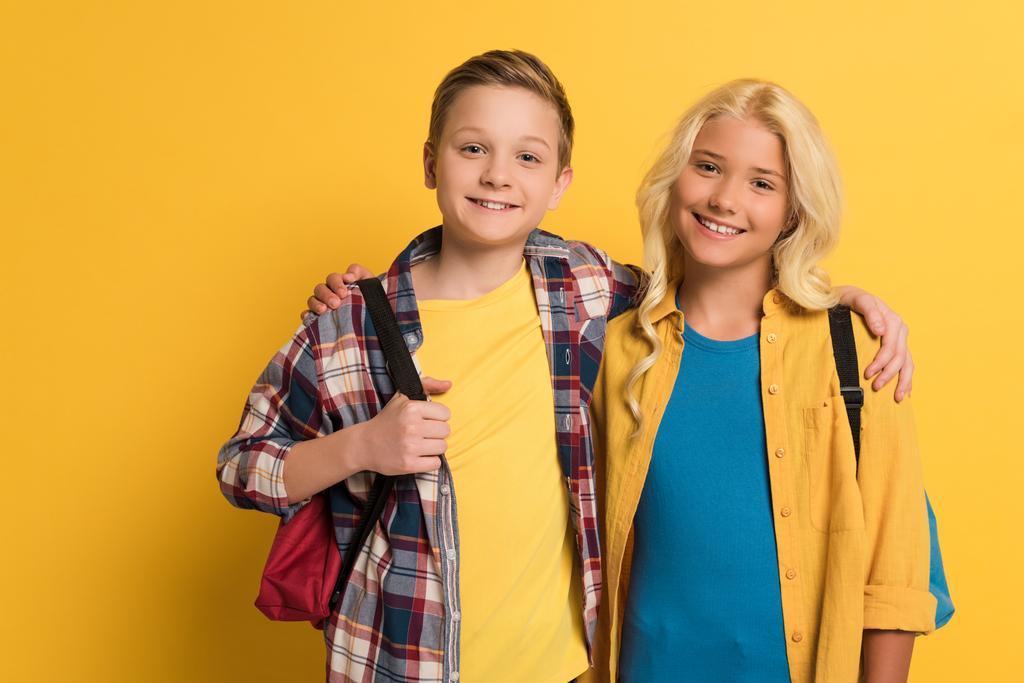 mosolygós iskolás gyerekek hátizsákkal ölelés és nézi kamera sárga háttér  - Fotó, kép