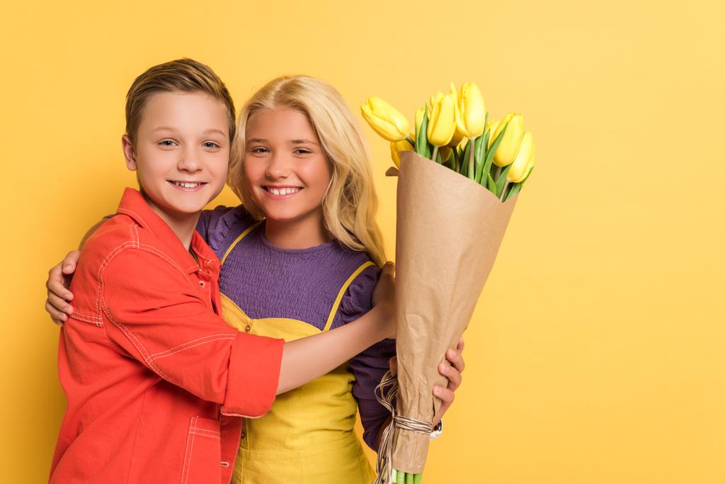 smiling boy hugging friend with bouquet on yellow background  - Photo, Image