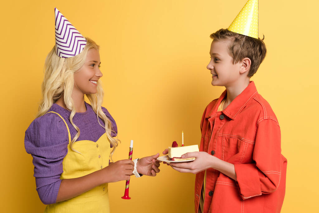 boczny widok chłopca dając talerz z tortem urodzinowym do uśmiechniętego przyjaciela z wieczkiem party na żółtym tle  - Zdjęcie, obraz