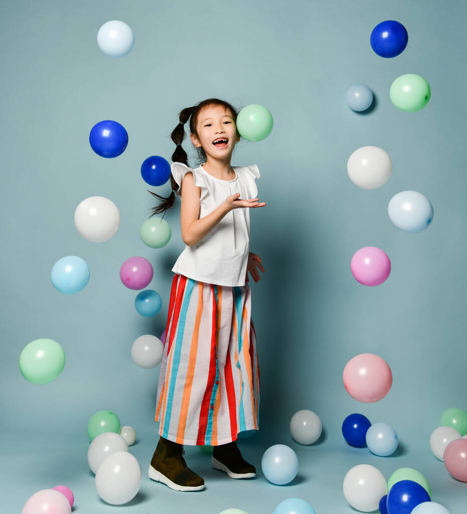 Сміється азіатська дівчинка в партійному одязі розважається в оточенні різнокольорових повітряних куль тримає руку з відкритою долонею
 - Фото, зображення