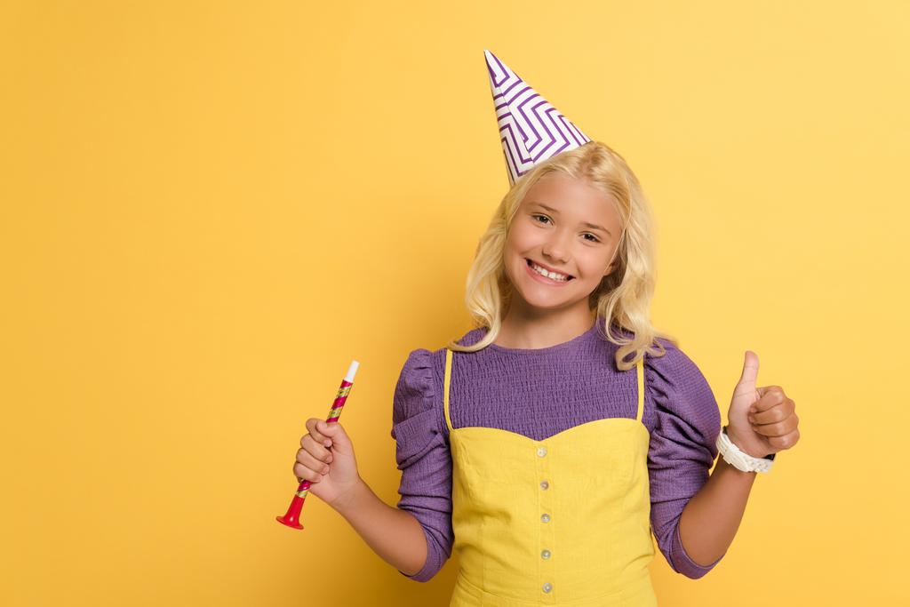 Lächelndes Kind mit Partymütze, Parteihorn in der Hand und wie auf gelbem Hintergrund  - Foto, Bild