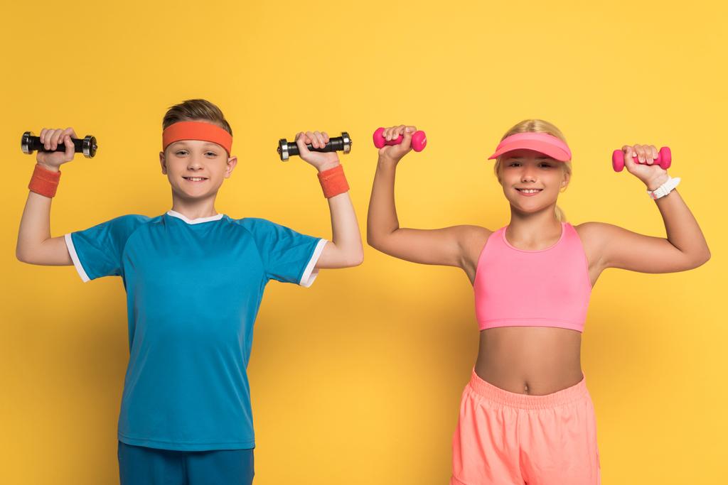 uśmiechnięte dzieci trenujące w odzieży sportowej z hantlami na żółtym tle  - Zdjęcie, obraz