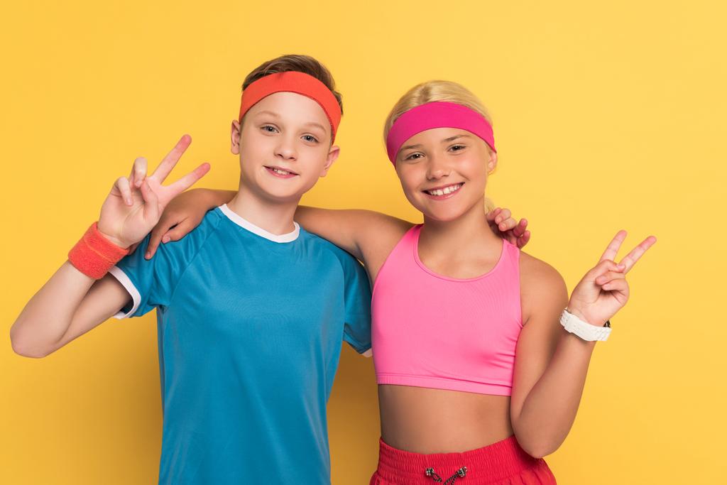 スポーツウェアを着た笑顔の子供たちが黄色の背景に平和のジェスチャーを示し  - 写真・画像