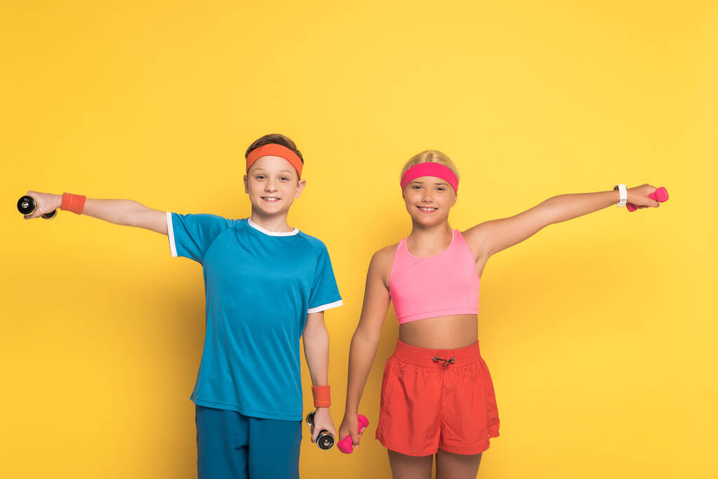 χαμογελαστά παιδιά σε αθλητικά ρούχα κατάρτισης με αλτήρες σε κίτρινο φόντο  - Φωτογραφία, εικόνα