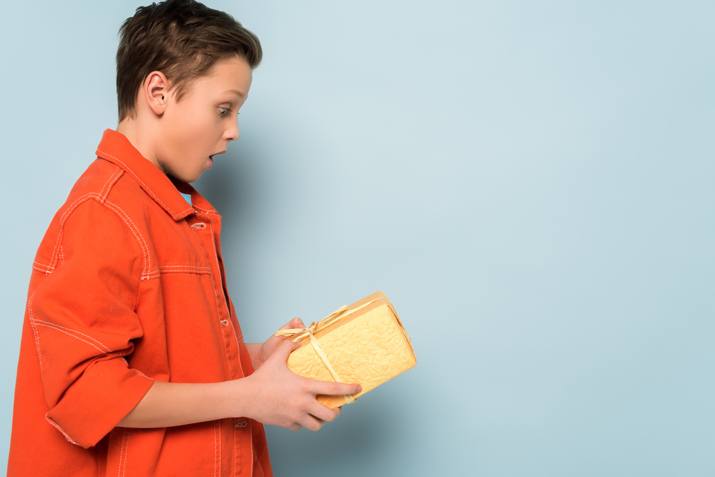 πλαϊνή άποψη του έκπληκτος παιδί κρατώντας κουτί δώρου σε μπλε φόντο  - Φωτογραφία, εικόνα