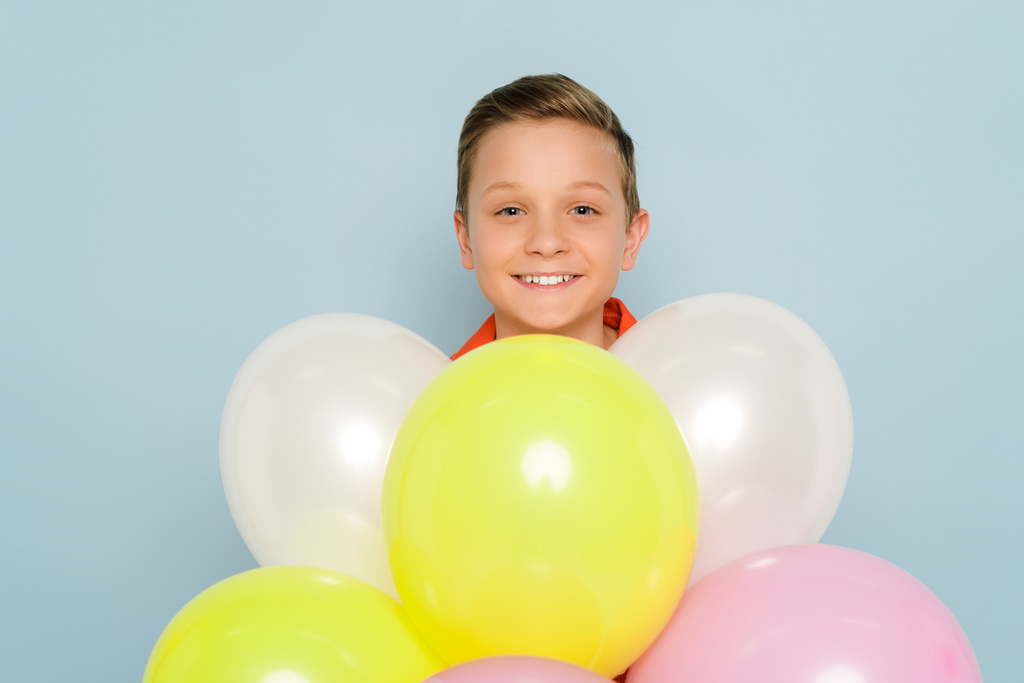 χαμογελαστό παιδί στέκεται κοντά μπαλόνια και κοιτάζοντας κάμερα σε μπλε φόντο  - Φωτογραφία, εικόνα