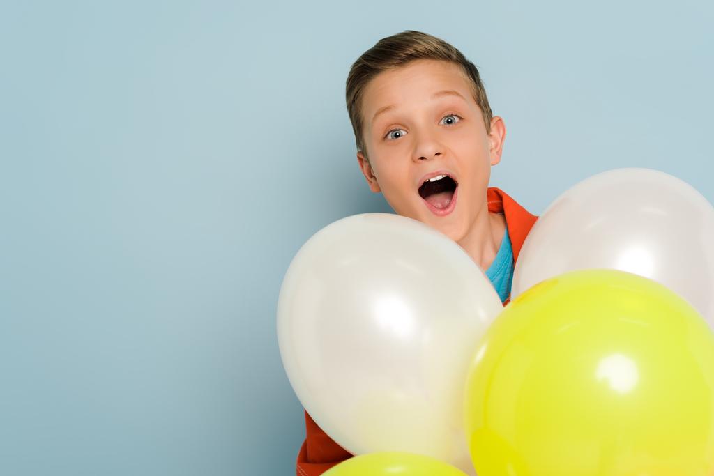Überraschtes Kind steht in der Nähe von Luftballons und blickt in die Kamera auf blauem Hintergrund  - Foto, Bild
