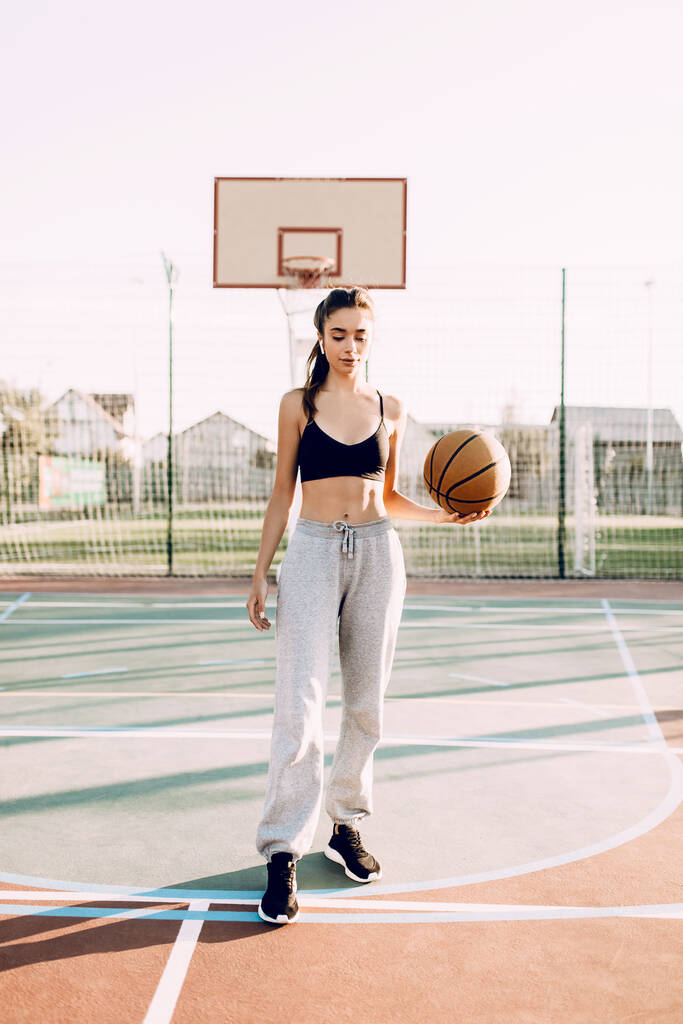 Hermosa joven atlética, en la parte superior y pantalones de chándal, jugando con la pelota en la cancha de baloncesto al aire libre
. - Foto, Imagen
