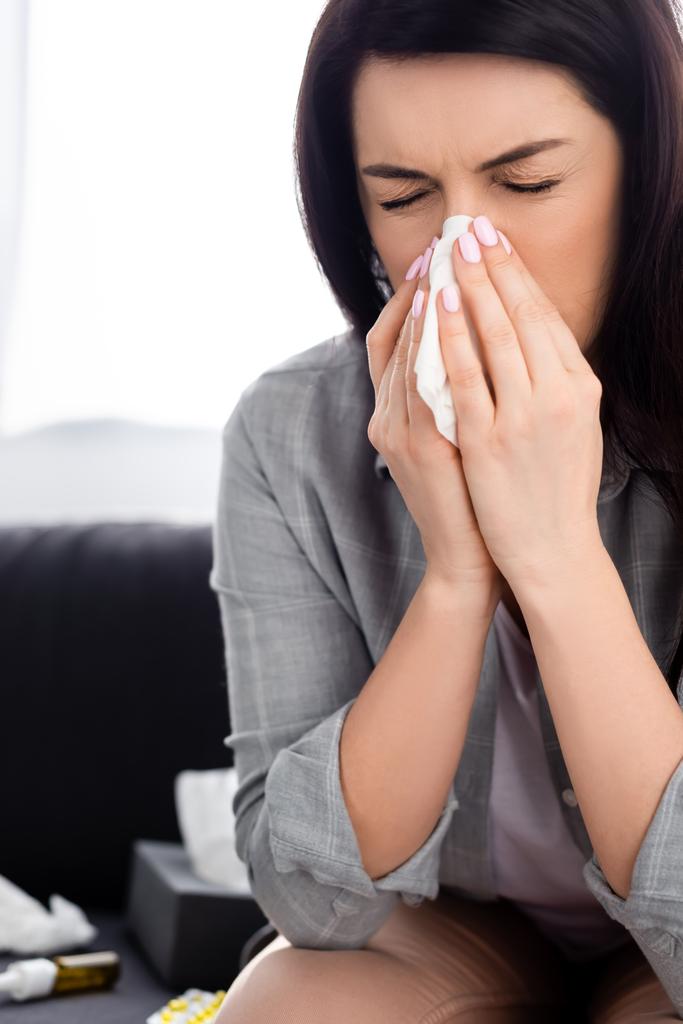 Allergische Frau mit geschlossenen Augen niest in Serviette, während sie auf dem Sofa sitzt  - Foto, Bild