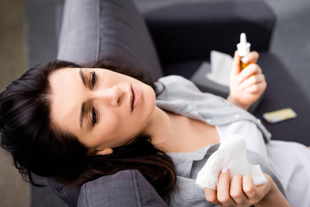 selective focus of allergic woman holding tissue and nasal spray while sitting on sofa  - Photo, Image