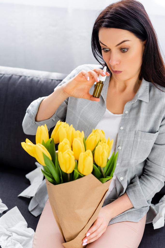 woman with pollen allergy sitting on sofa with flowers and holding nasal spray  - Photo, Image