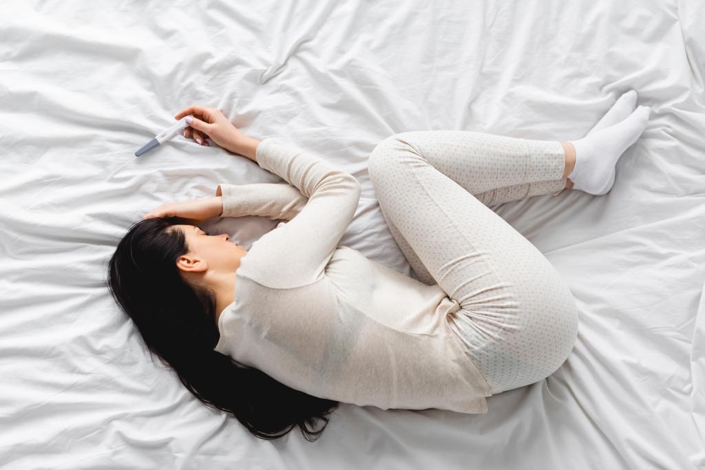 Ansicht einer depressiven Frau, die in der Nähe eines Schwangerschaftstests auf dem Bett liegt, mit negativem Ergebnis  - Foto, Bild