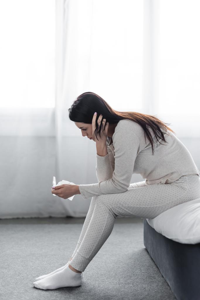 Depressive Frau sitzt auf Bett und schaut Schwangerschaftstest mit negativem Ergebnis an  - Foto, Bild