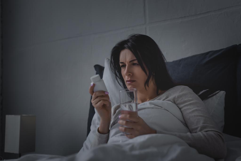 жінка з розладом сну тримає склянку води і пляшку зі снодійними таблетками в спальні
  - Фото, зображення