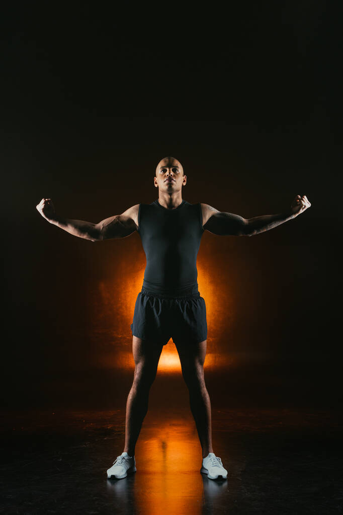 Guapo deportista fuerte en ropa deportiva mostrando bíceps. Luz naranja en el fondo
 - Foto, imagen