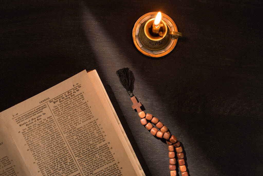 КИЕВ, УКРАИНА - 17 ЯНВАРЯ 2020: вид сверху на открытую Святую Библию с четками и свечами на темном фоне с солнечным светом
 - Фото, изображение
