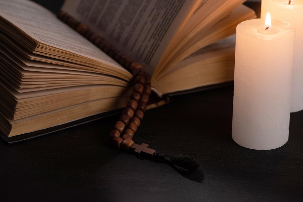 Santa Biblia con rosario sobre fondo negro oscuro con velas encendidas
 - Foto, Imagen