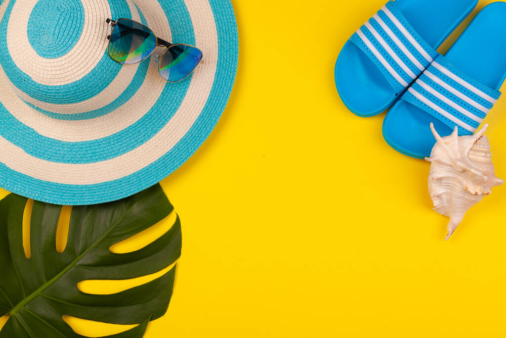 黄色の背景に青いビーチアクセサリーのトップビュー-サングラス、フリップフロップとストライプの帽子、シェルとモンステラの葉。ビーチの休日、海のツアー、暖かい晴れた夏の概念。広告スペース - 写真・画像