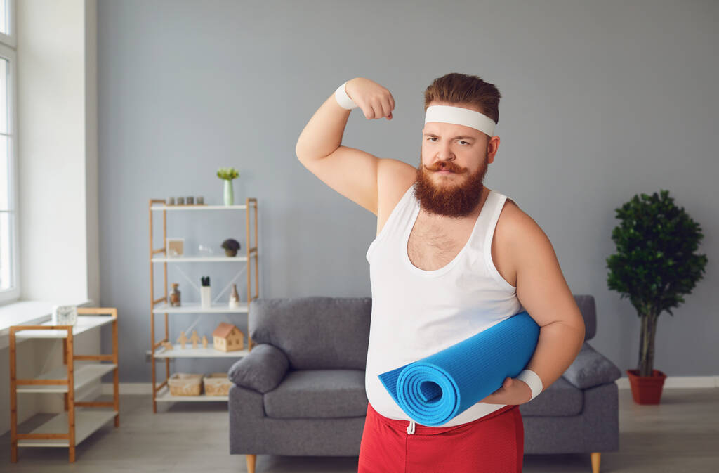 Αστείος χοντρός με αθλητικά δείχνει μύες να στέκονται στο δωμάτιο στο σπίτι. - Φωτογραφία, εικόνα