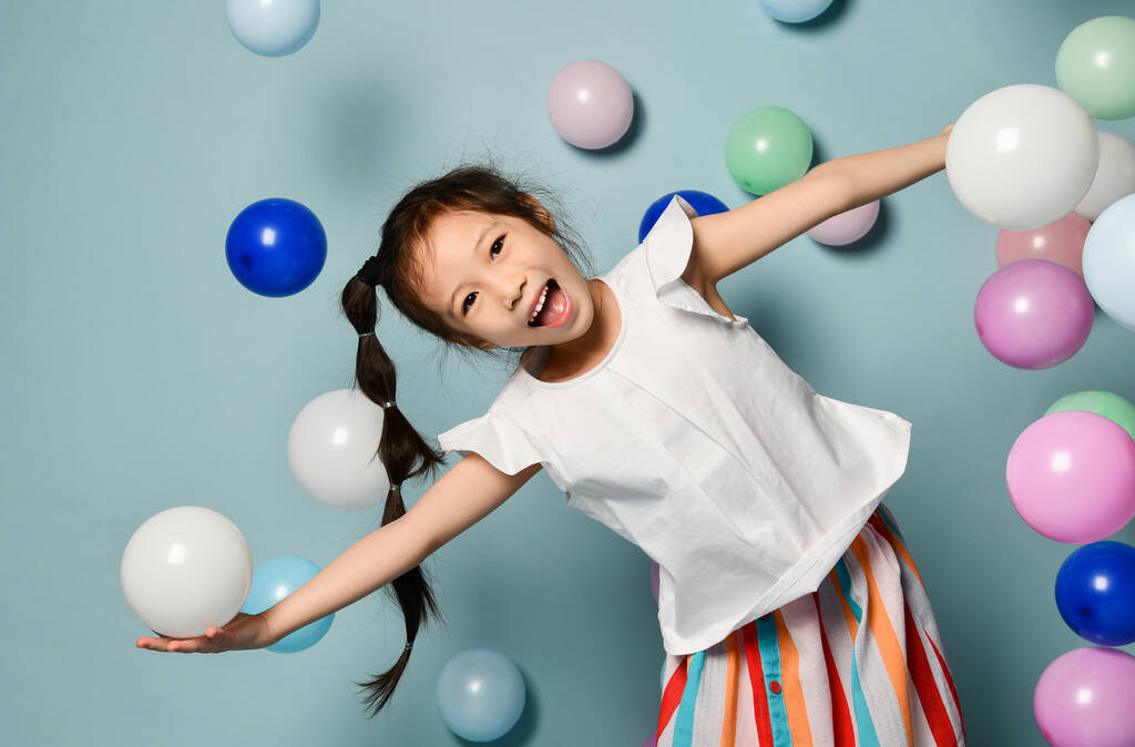 Iloinen aasialainen lapsi tyttö pitkä musta plait on hauskaa ympäröi värikäs ilmapalloja, levitä kädet
 - Valokuva, kuva