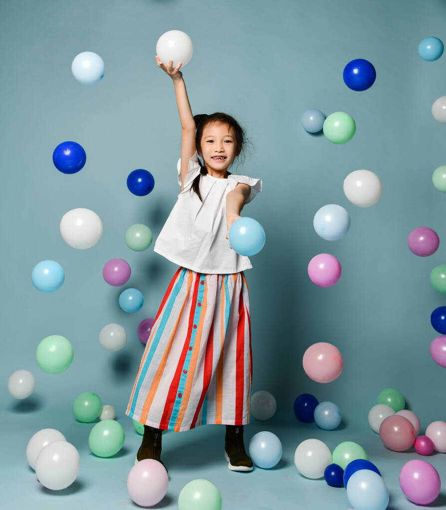 Onnellinen aasialainen lapsi tyttö tyylikäs raidallinen maxi hame on hauskaa näytelmiä ympäröi värikäs ilmapalloja tilalla oksentaa
 - Valokuva, kuva