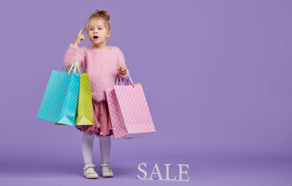 Sale of childrens products. Portrait of fun little child girl  on purple background holding shopping bags, package. Looking at the camera. Banner. Space for text.  - Photo, Image