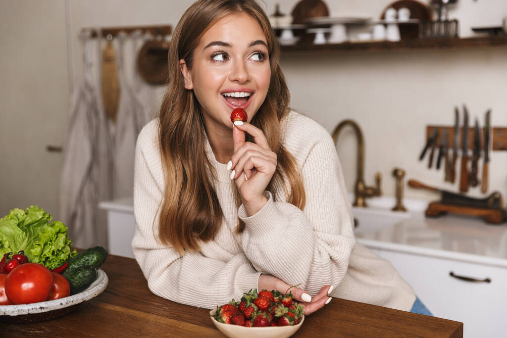 Εικόνα της ευτυχισμένης καυκάσιας γυναίκας που φοράει καθημερινά ρούχα τρώγοντας φράουλα και χαμογελώντας στην ζεστή κουζίνα - Φωτογραφία, εικόνα