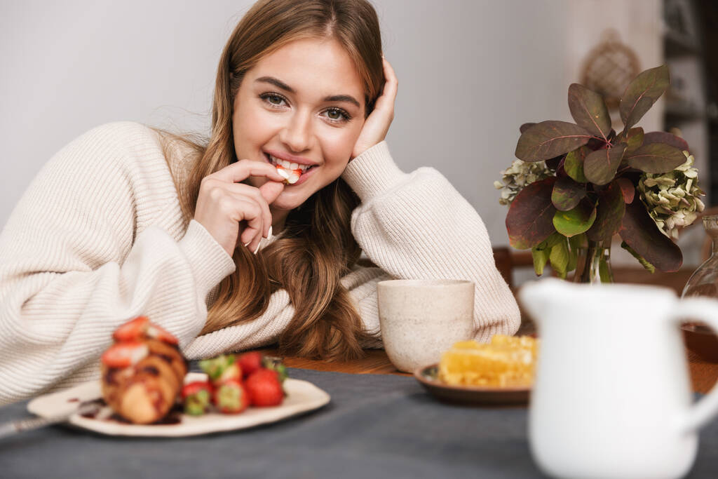 Obraz szczęśliwy biały kobieta noszenie casual ubrania jedzenie truskawki podczas śniadania w przytulnym pokoju - Zdjęcie, obraz