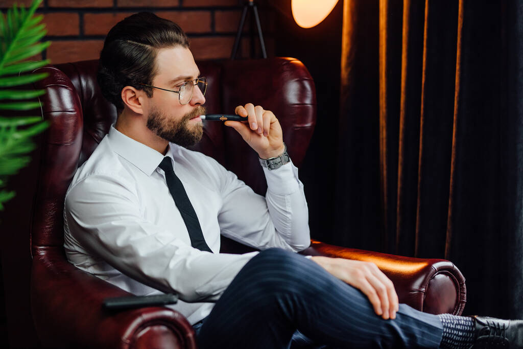 Zakenman rusten en ontspannen terwijl zitten op de bank in luxe kamer. Knappe man die sigaren rookt of iqos. - Foto, afbeelding