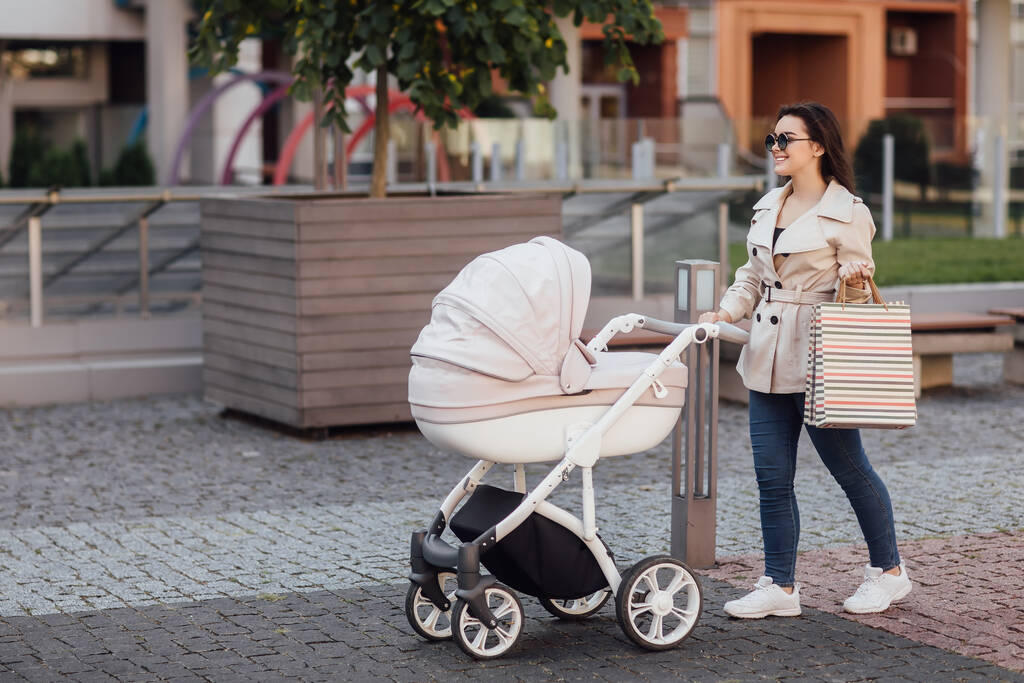 Hora de ir de compras. Retrato de madre bonita con paquete de compras caminando con carro de bebé en el centro de la ciudad europea
. - Foto, imagen
