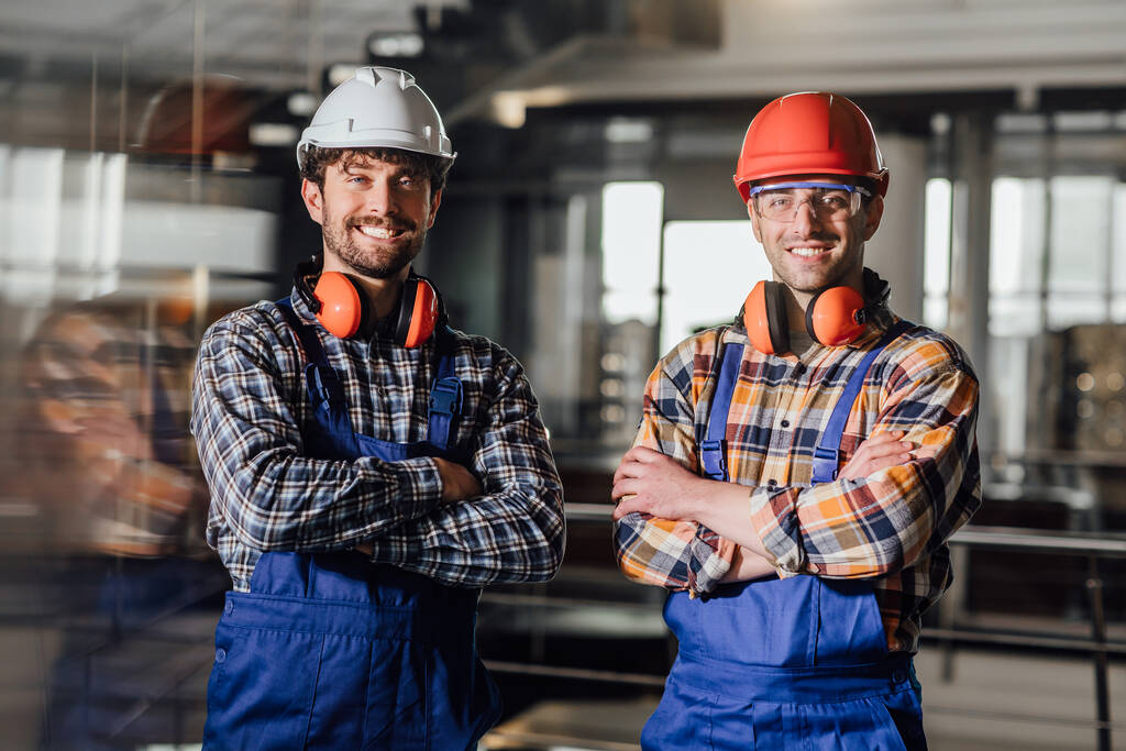 Dos jóvenes constructores en uniforme de trabajo y cascos cruzados de brazos
. - Foto, Imagen