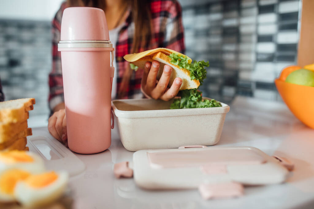 Photo rapprochée, jeune fille tient son sandwich complet sain sur sa boîte à lunch et thermos à la maison cuisine
. - Photo, image