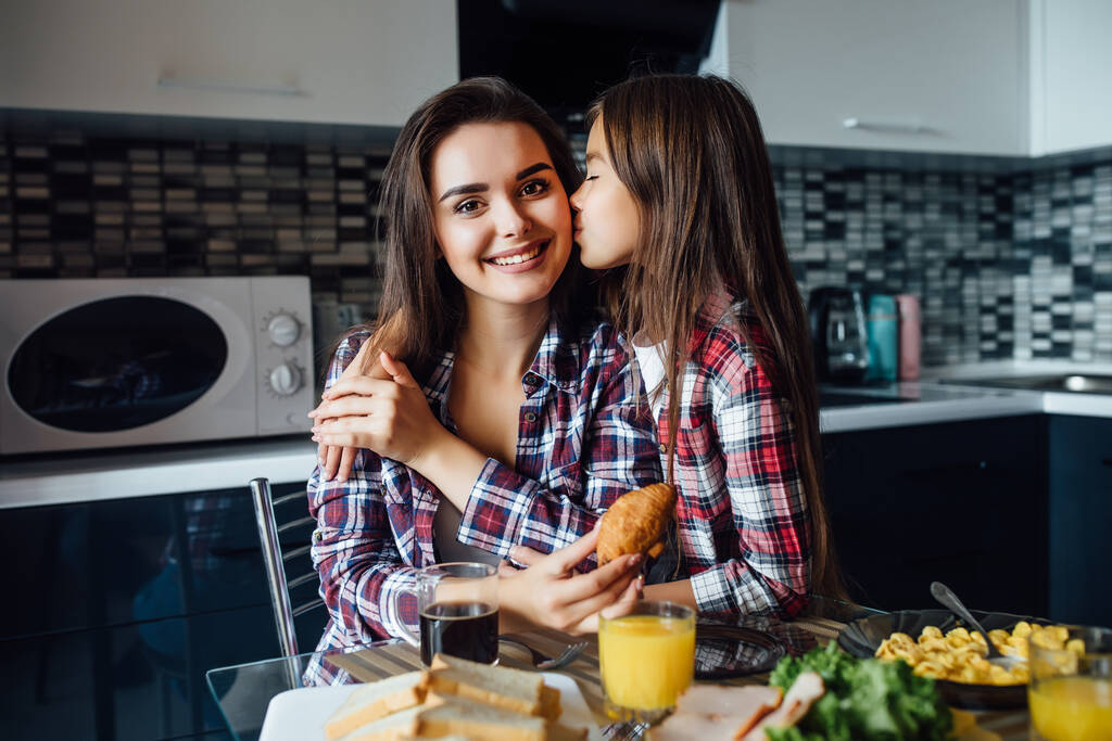 Adorable girl is having an healthy snack with cookies and milk with her mother, while kiss on the cheek. - Photo, Image