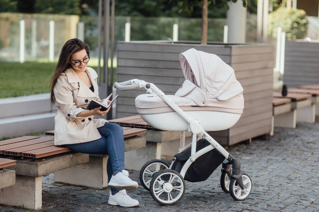 hymyilevä äiti käsittelyssä kirja penkillä lähellä vauvan rattaat puistossa
. - Valokuva, kuva