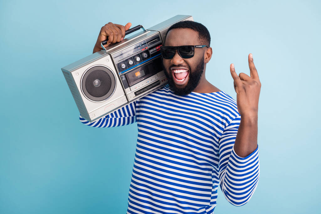 Hard rock. Photo of funny dark skin guy hold retro cassette recorder on shoulder showing metal horns hand wear sun glasses striped sailor shirt isolated blue color background - Photo, Image