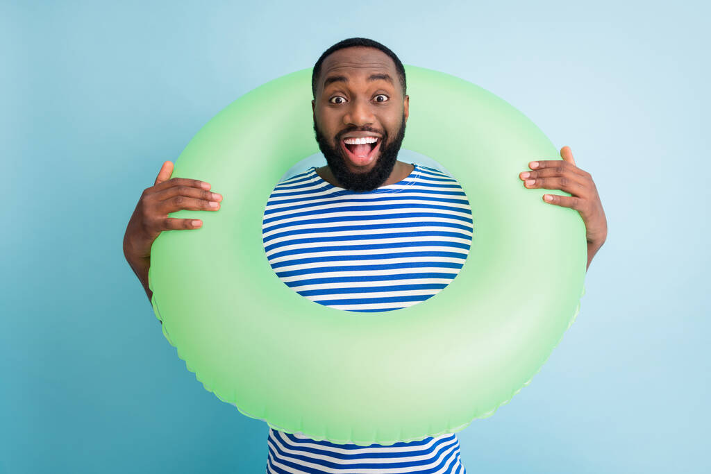 Photo of funky dark skin guy hold green life buoy ready swim ocean sea traveler see sale banner advert enjoy shopping rest abroad wear striped sailor shirt isolated blue color background - Zdjęcie, obraz