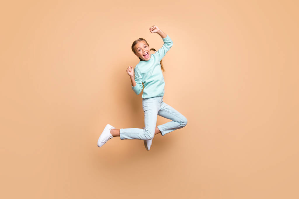 Full body profile photo of beautiful funny little lady jumping high up celebrating winning raise fists wear blue sweater jeans footwear isolated beige color background - 写真・画像