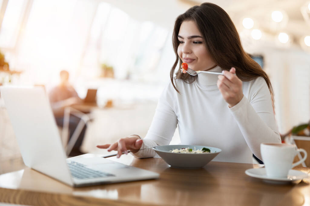 joven mujer hermosa positiva trabaja en su computadora portátil sosteniendo tenedor con tomate mientras come ensalada en la cafetería durante la hora del almuerzo, concepto multitarea
. - Foto, imagen