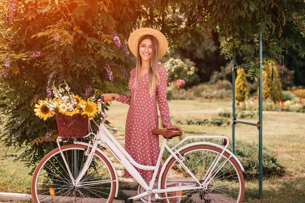 Giovane donna soddisfatta in cappello di paglia e bicchieri con bicicletta in giardino estivo
 - Foto, immagini