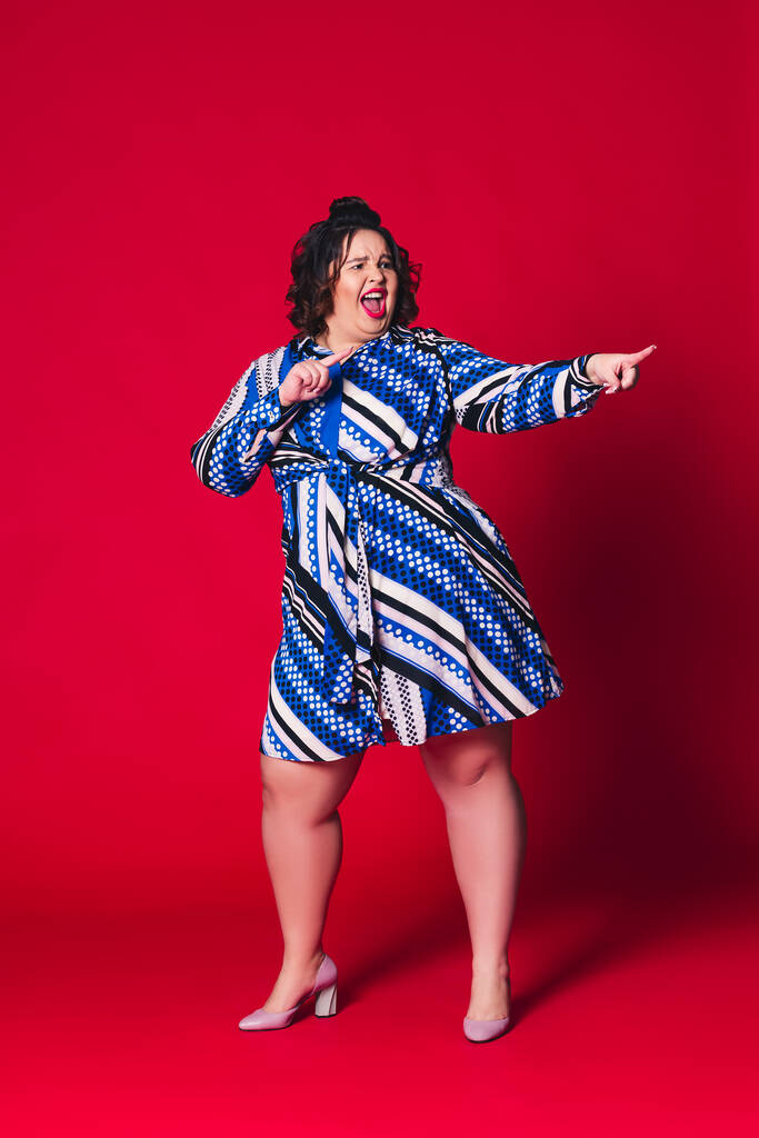 Glückliches Pize-Size-Model im blauen Kleid, dicke Frau tanzt auf rotem Hintergrund, körperbetontes Konzept - Foto, Bild