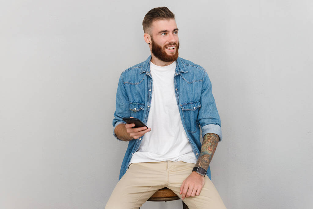 Портрет красивого усміхненого молодого чоловіка в повсякденному одязі, що сидить ізольовано на сірому фоні, використовуючи мобільний телефон
 - Фото, зображення