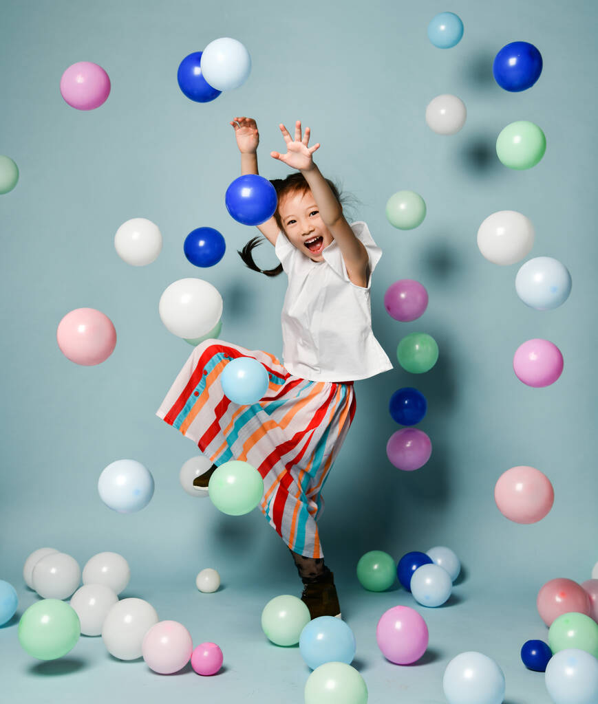 Happy asijské dítě dívka ve stylové pruhované maxi sukně má zábavu skoky skákání tance obklopené barevné vzduchové balónky - Fotografie, Obrázek