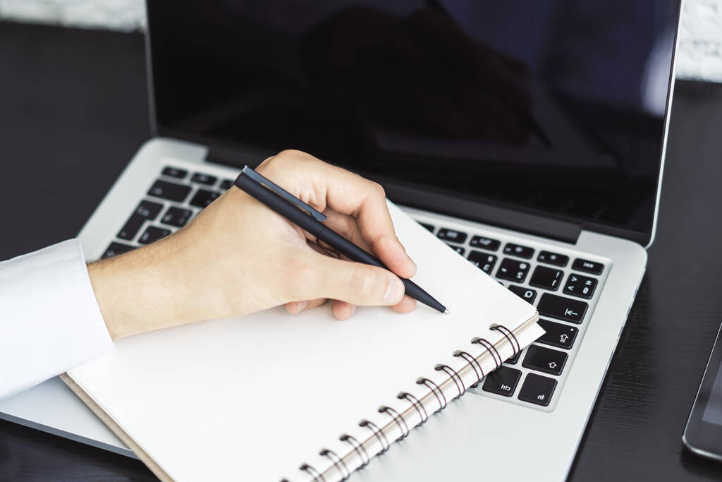 O homem escreve com uma caneta no bloco de notas no teclado do laptop em um escritório ensolarado, conceito de negócios e educação. Fechar
 - Foto, Imagem